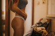 © qunica.com - Young woman wearing underwear stands in a softly lit room, holding her stomach. The intimate portrait captures a quiet moment in a beauty center setting.
