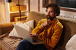 © Stockphotodirectors - A man in a cozy sweater is sitting on a couch, focused on his laptop in a warm living room. Soft lighting creates a relaxed atmosphere as he ponders his work.