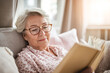 © Kowit - An elderly woman wearing glasses relaxes on a couch, reading a book in a cozy, sunlit room.