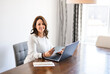 © Louis-Paul Photo - Senior woman at home kitchen table feeling goodd while looking at multiple bills working on her laptop