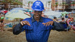 © Krakenimages.com - Man in blue hardhat and coveralls smiling and gesturing with both hands on a crowded beach with umbrellas and sand; work pride.