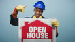 © Krakenimages.com - Man in blue hardhat points finger at red open house sign while wearing yellow gloves in studio; confidence promotion.