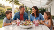 © Muhammad - Happy family eating healthy lunch on outdoor patio, smiling parents and children sharing meal together, summer garden dining with fresh salad and smoothies