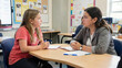 © Luisa - An indoor classroom scene where a young girl is talking to a teacher while the educator takes notes in a book.