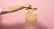 © fitria - A person's arm holding a brown paper lunch bag against a pink background.