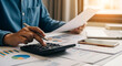 © fitria - A person using a calculator and reviewing financial documents at a desk with a laptop and smartphone.