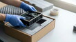 © CK Studio - Hands in blue gloves carefully place an electronic device into protective foam packaging within a cardboard box for shipping.