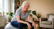 © Eve - Happy senior woman sitting on fitness ball at home. Smiling elderly female in grey t-shirt exercising for wellness and healthy lifestyle. Active retirement and physical therapy for seniors.