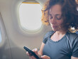 © Юлия Завалишина - Passenger woman is flying in plane. Girl using phone sitting by airplane window. Traveling female with smartphone inside plane enjoying flight. Woman using wifi connection on board. Traveling girl