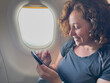 © Юлия Завалишина - Passenger woman is flying in plane. Girl using phone sitting by airplane window. Traveling female with smartphone inside plane enjoying flight. Woman using wifi connection on board. Traveling girl