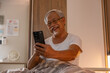 © 220 Selfmade studio - Elderly Asian man happily using smartphone in bed smiling with joy modern technology connection senior lifestyle communication resting relaxing indoor