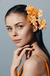 © VICHIZH - Beautiful young woman with glowing skin and orange flowers in her hair, wearing an orange outfit, showcasing a serene expression against a soft blue background.