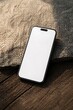 © muhagraph - A modern smartphone with a blank white screen rests on a textured wooden surface next to a large, rough stone.