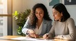 © Fathiyah - Two diverse women collaborating on a project at a desk, smiling and pointing at documents
