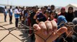 © Nataly - Displaced individuals waiting in line behind wire fence at refugee camp with humanitarian workers providing assistance.
