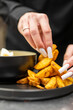 © pavel siamionov - Close-up of a person with long white manicured nails picking up seasoned, golden-brown potato wedges from a dark plate next to a black bowl of dipping sauce.