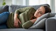 © olga_demina - A woman experiencing stomach pain lies on a couch in a cozy living room, clutching her stomach in discomfort.