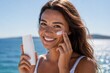 © mtmakos - Beautiful young woman applying sunscreen cream on her cheek while holding a blank cosmetic bottle. Shot at the beach with bright sunlight and natural skin texture. Perfect for skincare, UV protection,