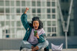 © carballo - A euphoric young woman with curly hair raises her fist in a victory gesture while checking her phone, sitting cross-legged in front of a modern glass building: energy, freedom, and digital triumph