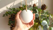 © abufatir - Hand holding a white ceramic Christmas ornament with pine cones and greenery.