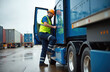 © Viktor - Man driver wearing hard hat, safety vest climbs into blue truck cab. Ready for road trip with cargo containers visible in background. Logistics industry professional begins work. He prepares to drive.