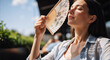 © NeuroCake - Young woman cooling her face with a hand fan on a sunny day. Female relaxing outside during summer heatwave