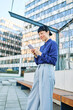 © Lumos sp - Portrait of a young asian japanese businesswoman woman using a smartphone mobile phone walking down the street, surrounded by moder corporate office buildings