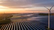 © picbox - Rows of solar panels and several wind turbines are spread across a green hilltop, illuminated by the warm glow of a sunset. The stunning landscape highlights the commitment to sustainable energy