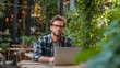 © vanilnilnilla - A man works on his laptop in an outdoor garden cafe  long title A man wearing casual attire sits at a table in an urban garden cafe using his laptop