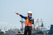 © VStudio - Woman wears a white hard hat and orange safety vest. She points with a pen while holding a tablet. woman engineer communicating via radio while inspecting maritime logistics at an industrial harbor.