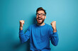 © miss irine - Bearded man in glasses wears blue sweater, fists raised in joy. He shouts in triumph, eyes closed, celebrating a win. Success is achieved, pure happiness shown.