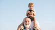 © Elena - Three generations of smiling men with a grandfather carrying his adult son and young grandson on his shoulders against a clear blue sky