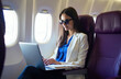 © Vadym - Young woman wearing sunglasses works on laptop inside airplane cabin. She sits by window, typing on computer and holding pen. Pleasant flight journey with work during trip.