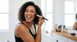 © ibhonk - Joyful mixed ethnicity adult woman with dark curly hair applying makeup brush to cheek in modern, brightly lit bathroom.