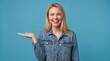 © ibhonk - Cheerful young adult woman with long blonde hair holds her open hand out presenting space on a solid blue  backdrop.