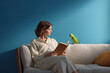 © Jackson Photography - Young woman reading a book on a sofa with a green parrot beside her, set against a bold blue background in a modern interior.