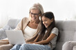 © fizkes - Little 7s girl and older 55s granny relaxing on sofa with laptop, watching comedy movie, laugh, feel happy. Multigenerational relatives spend free time on internet, giggling while enjoy new videovlog