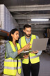 © fizkes - Warehouse employees standing next to storage racks, using laptop, checking inventory records, updating storehouse system. Logistics workflow, supply chain management, distribution center operations