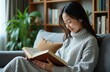 © Pete - Young Asian woman in comfy sweater reads book on sofa. She enjoys quiet indoor time by bookshelf, focusing on literature and relaxation. Casual home leisure and learning.