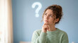 © Abelorz - Confused young woman with hand on chin looking up at a glowing neon question mark icon above her head against a blue wall background thinking about a problem or making a difficult decision