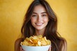 © btiger - person with long brown hair and blurred face holding a white bowl overflowing with potato chips against a warm yellow background, inviting and cheerful