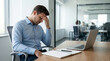 © Jai Izzam - Stressed overworked businessman with headache holding head in hand at wooden desk with laptop in bright modern corporate office