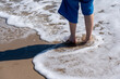 © Sergej - A refreshing wave of sea foam surrounds a person's feet on a sunny beach, captured in sharp detail.