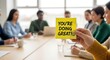 © MopoWe - Positive feedback sticky note held by a hand in a bright office during a team meeting with beautiful bokeh. This is ideal for employee engagement articles and leadership training presentations.