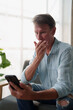 © Tj - Mature man reacting with shock and disbelief, holding his hand over his mouth while looking at his smartphone, processing unexpected news or a surprising message at home