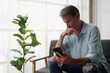 © Tj - Mature man looking down at cell phone screen, hand on mouth, experiencing concern or bad news, contemplating a serious issue at home