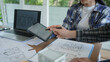 © NanSan - Architects reviewing house floor plans at desk, laptop and tablet in bright office, collaborating on home design project with blueprints and documents.