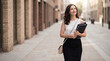 © Tati - A woman walks along a city street holding folders in both hands. She has a confident expression and is dressed in a simple outfit. The sun shines brightly, casting shadows