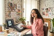 © Asier - Young woman talking on phone and designing on a digital tablet at her home studio