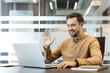 © Liubomir - Smiling young man making a video call on a laptop, waving his hand, actively participating in an online meeting or virtual conference from a modern office environment